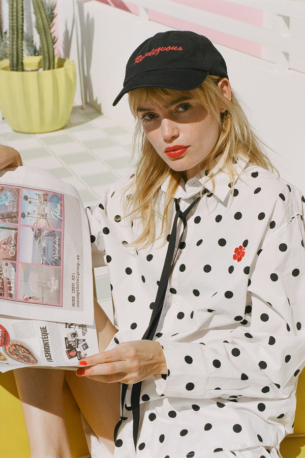 Woman wearing a black and white polka dot jacket with a black cap, holding a newspaper.