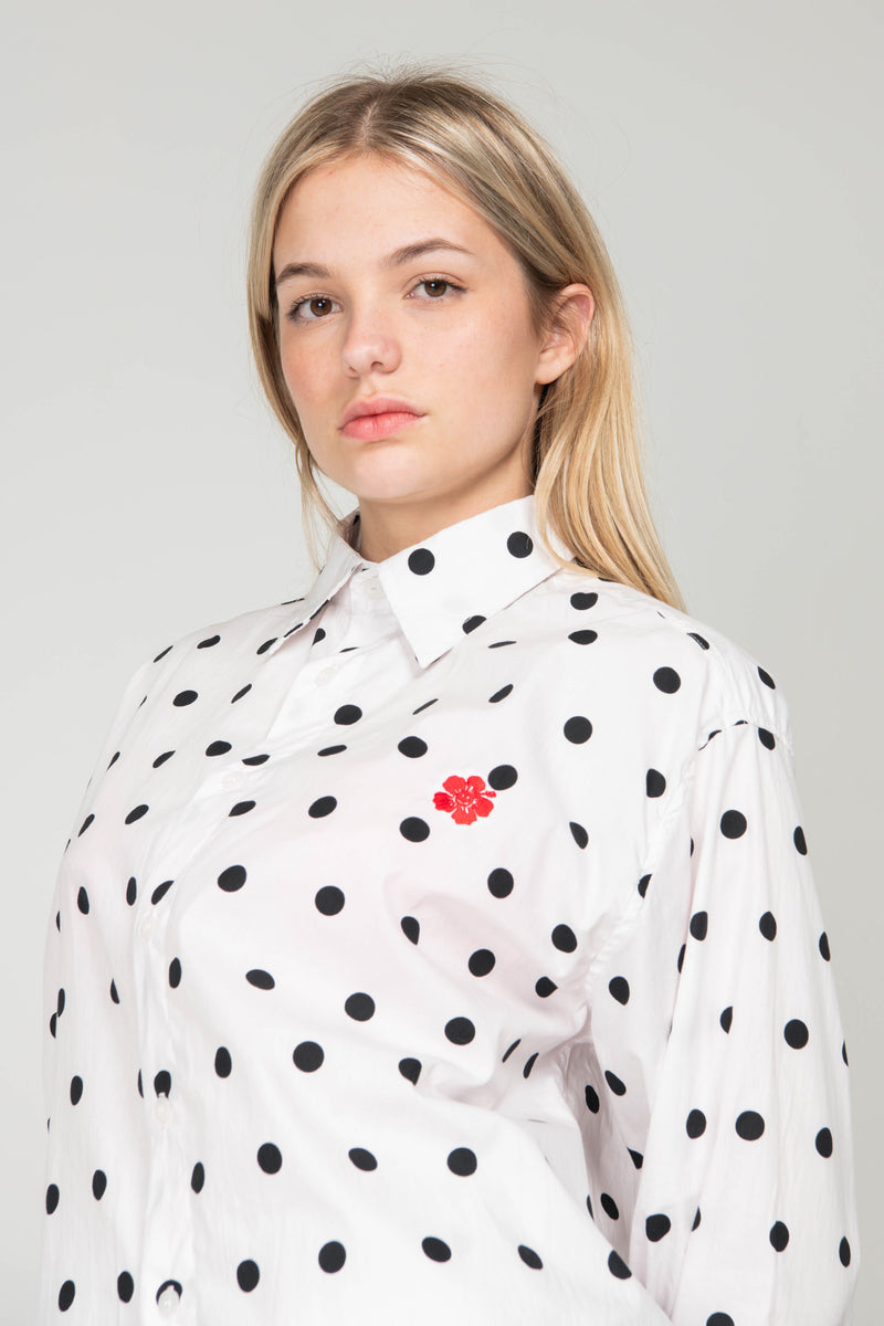Person wearing a white shirt with black polka dots and a red flower emblem on a plain background