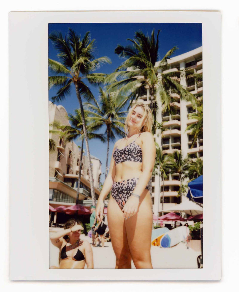 Woman in a bikini standing in front of palm trees and a hotel.