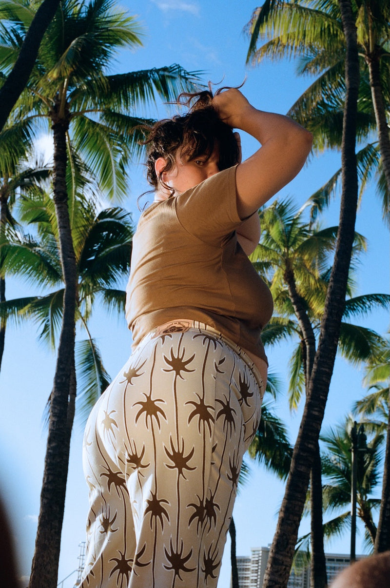 Person in a beige shirt and patterned pants standing among palm trees with a clear blue sky.
