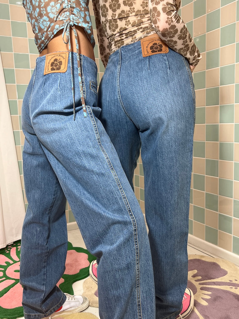 Person wearing blue jeans with a patterned top and belt, standing against a tiled wall.