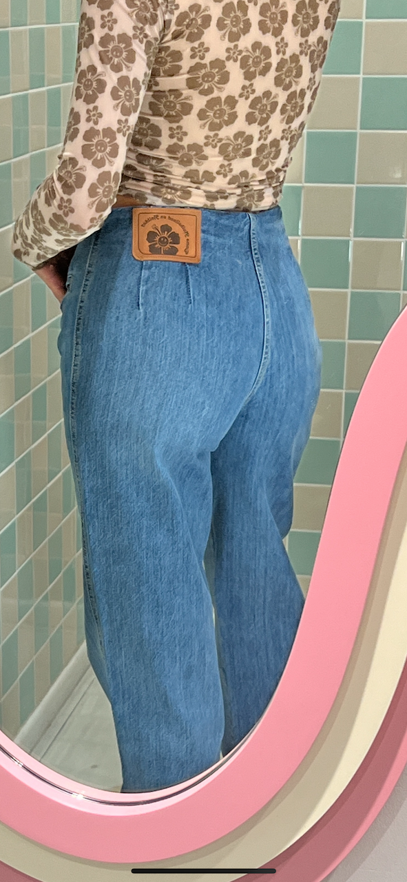 Person wearing blue jeans and a patterned shirt with a belt, standing in front of a tiled wall.