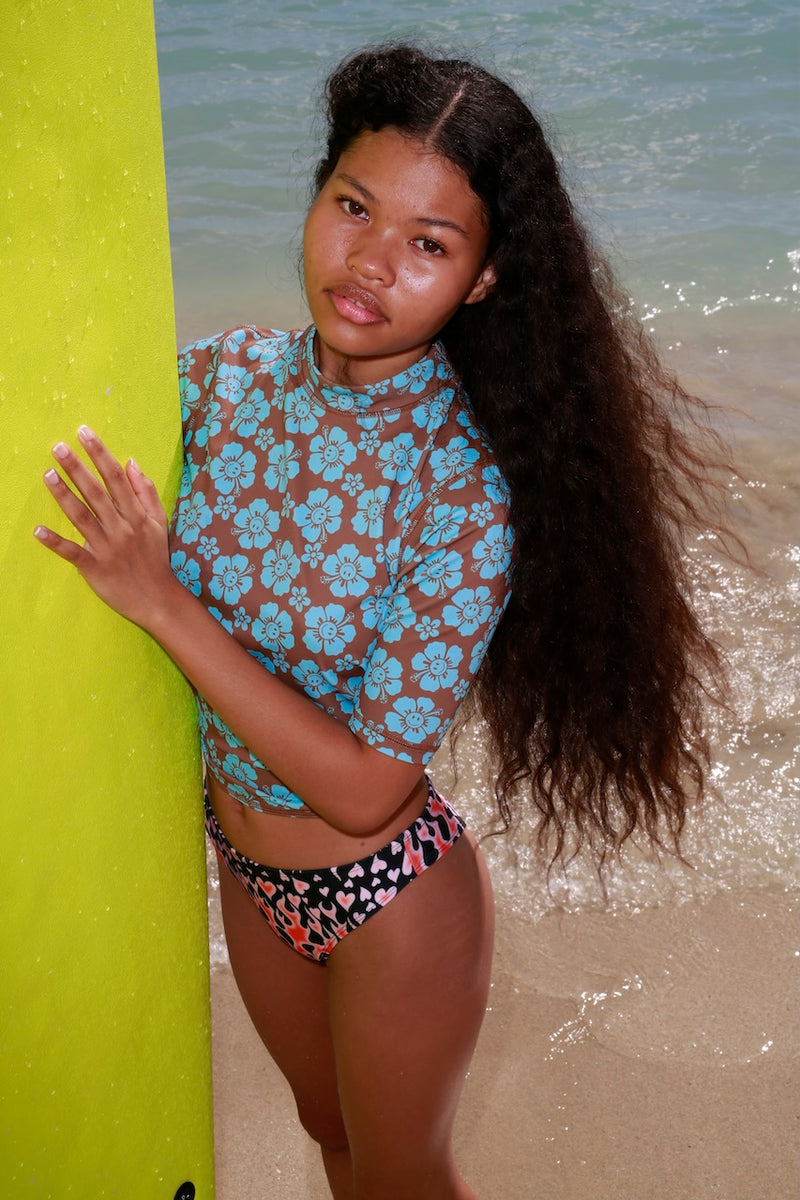 Woman in a floral top and patterned shorts standing next to a yellow surfboard on a beach.