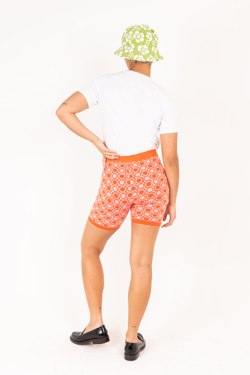 Person wearing orange patterned shorts and a white shirt with a green floral hat on a white background