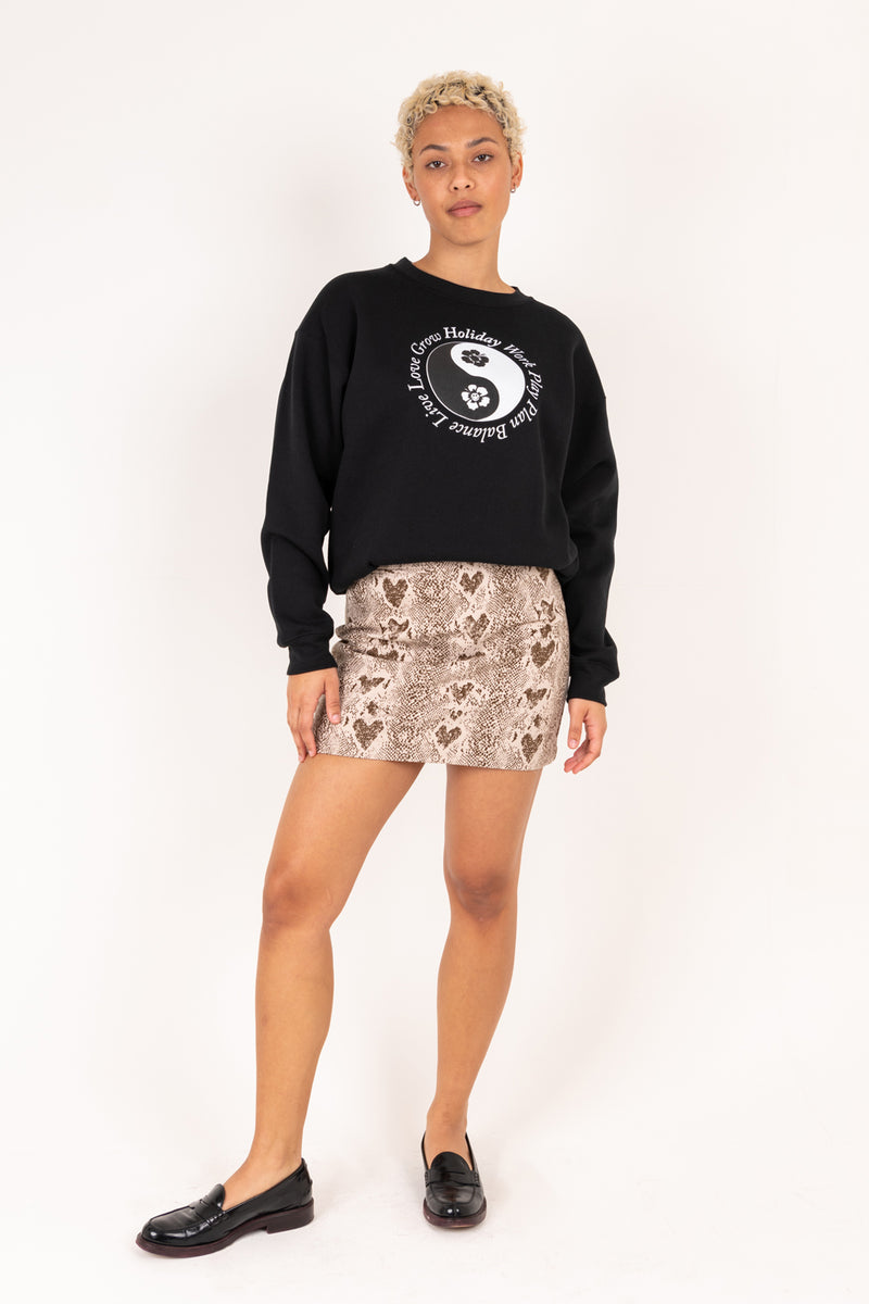 Person wearing a black sweatshirt with a white logo and a patterned skirt on a white background