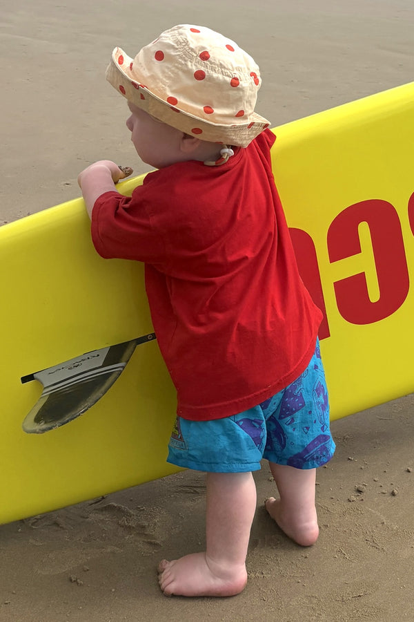 Child in red shirt and blue shorts standing next to a yellow surfboard on a sandy beach.