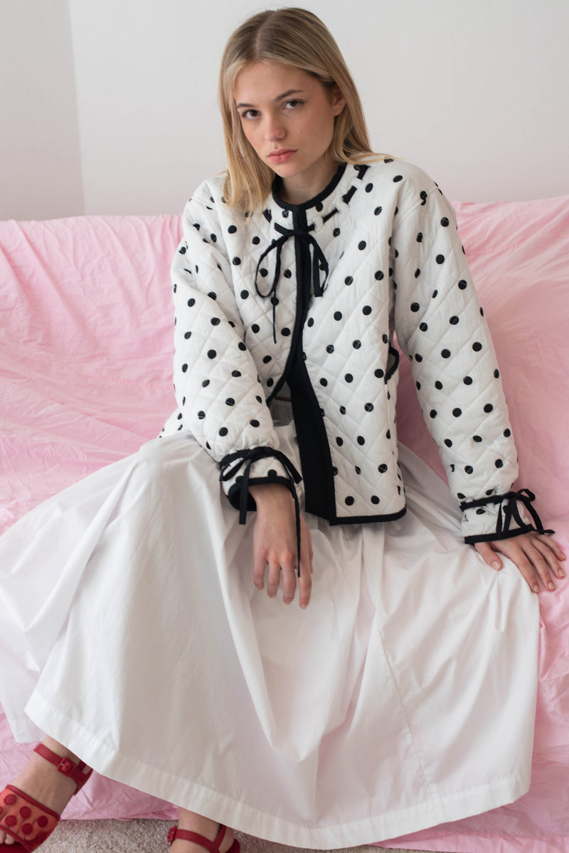 Woman wearing a polka dot jacket and white skirt sitting on a pink couch.