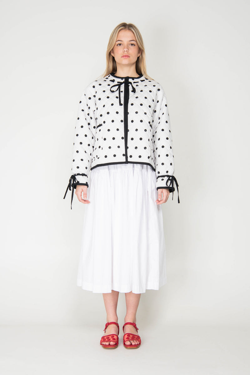 Woman wearing a black and white polka dot reversible jacket with a white skirt on a white background