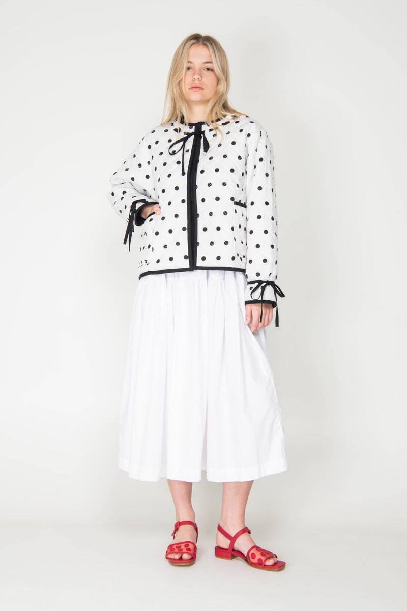 Woman wearing a black and white polka dot reversible jacket with a white skirt on a white background