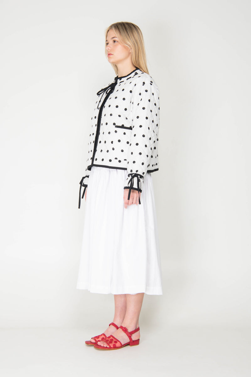 Woman wearing a black and white polka dot reversible jacket with a white skirt on a white background
