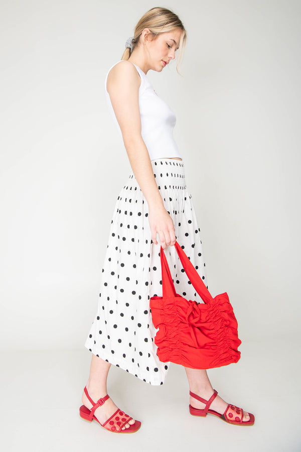 Woman holding a red bag wearing a white top and polka dot skirt on a plain background